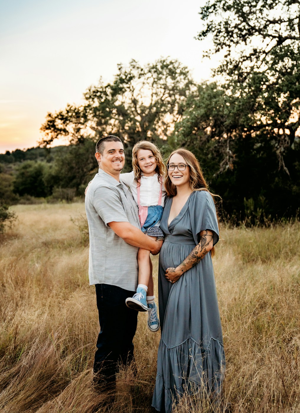 Family of three standing together in a grassy meadow at sunset